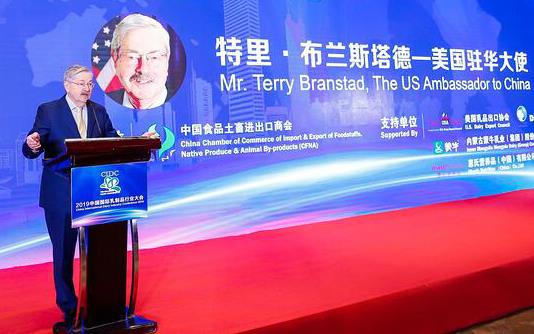 Older white man wearing business suit stands at podium with large poster welcoming Terry Branstad, US Ambassador to China