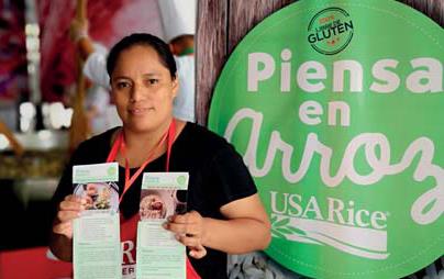 Woman wearing red apron holds recipe cards in both hands, standing in front of promotional banner that says "Piensa en Arroz"