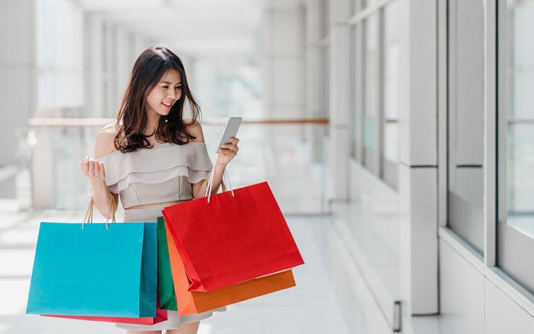 Young Asian woman walking in a mall holds multiple, colorful shopping bags and a cellphone