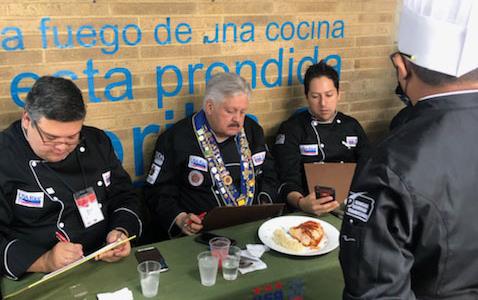 Panel of three judges wearing black chefs jackets sit behind table with green cloth and a dish of food