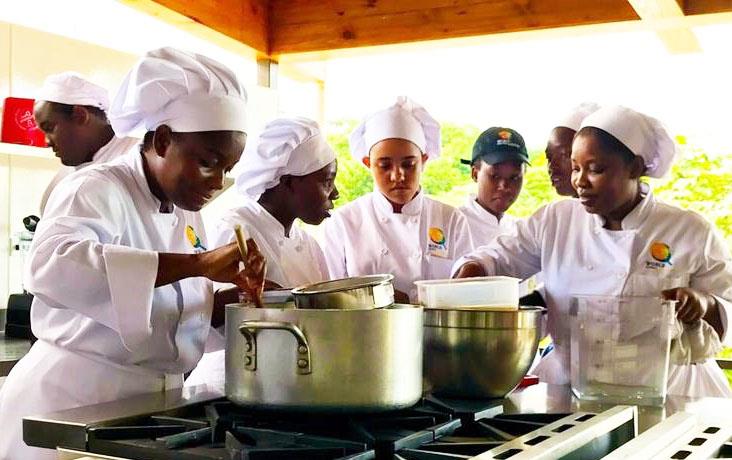 Student chefs wearing white chef jackets and toques gather around pots on stove