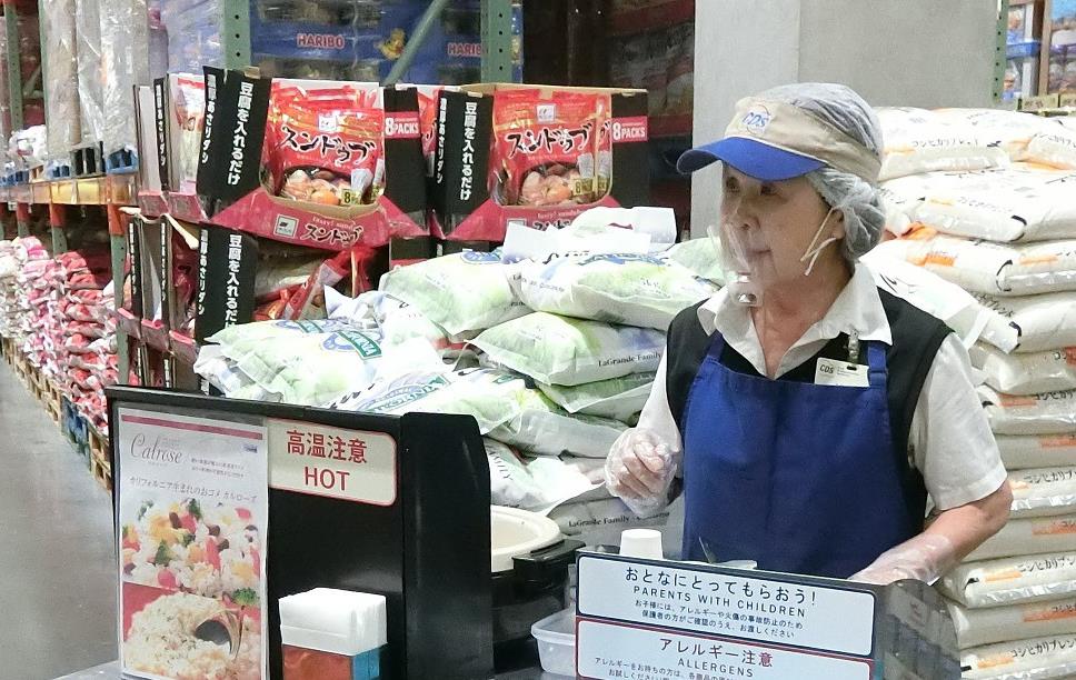 Woman wearing blue apron, hairnet, and visor stands behind metal cart handing out rice samples in paper cups