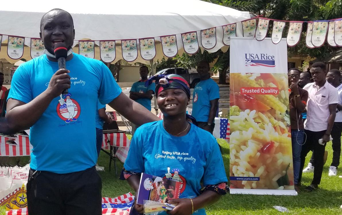 Man and woman wearing blue Ricky Rice t-shirts at a Ghana-promotion-seminar outside, banners and signage in background