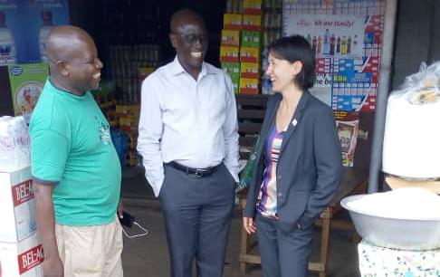 White woman wearing business suit stands with two casually dressed black men in street market stall