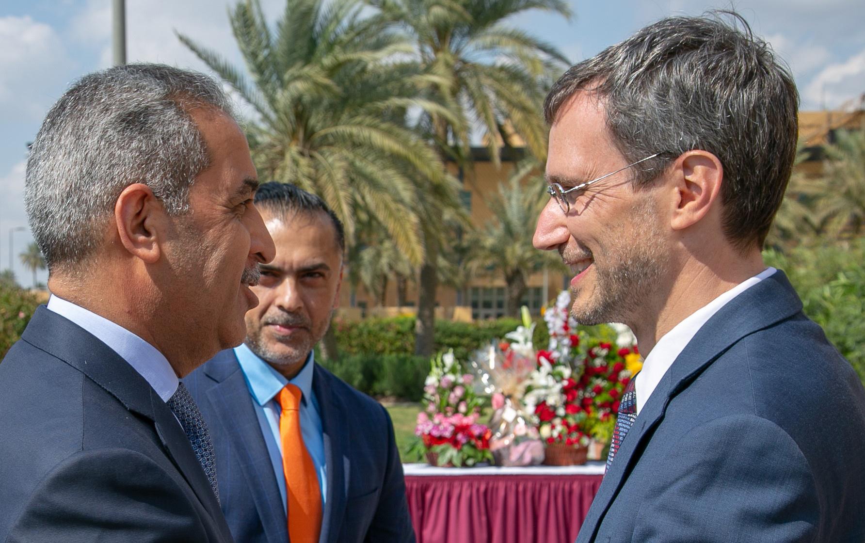 Three men in blue business suits stand together outside near palm trees and table full of flowers