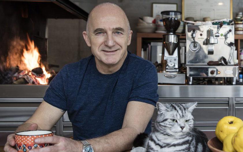 Bald white man wearing dark shirt, holds a cup in his hands, gray tabby cat and bowl of golden pears next to him, fire and kitchen equipment in background