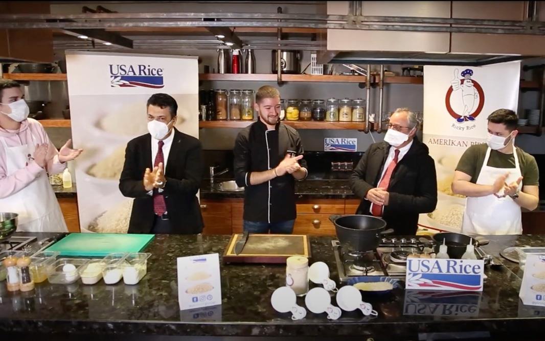 Group of men, some wearing suits, others wearing chefs jackets, stand behind cooking station 