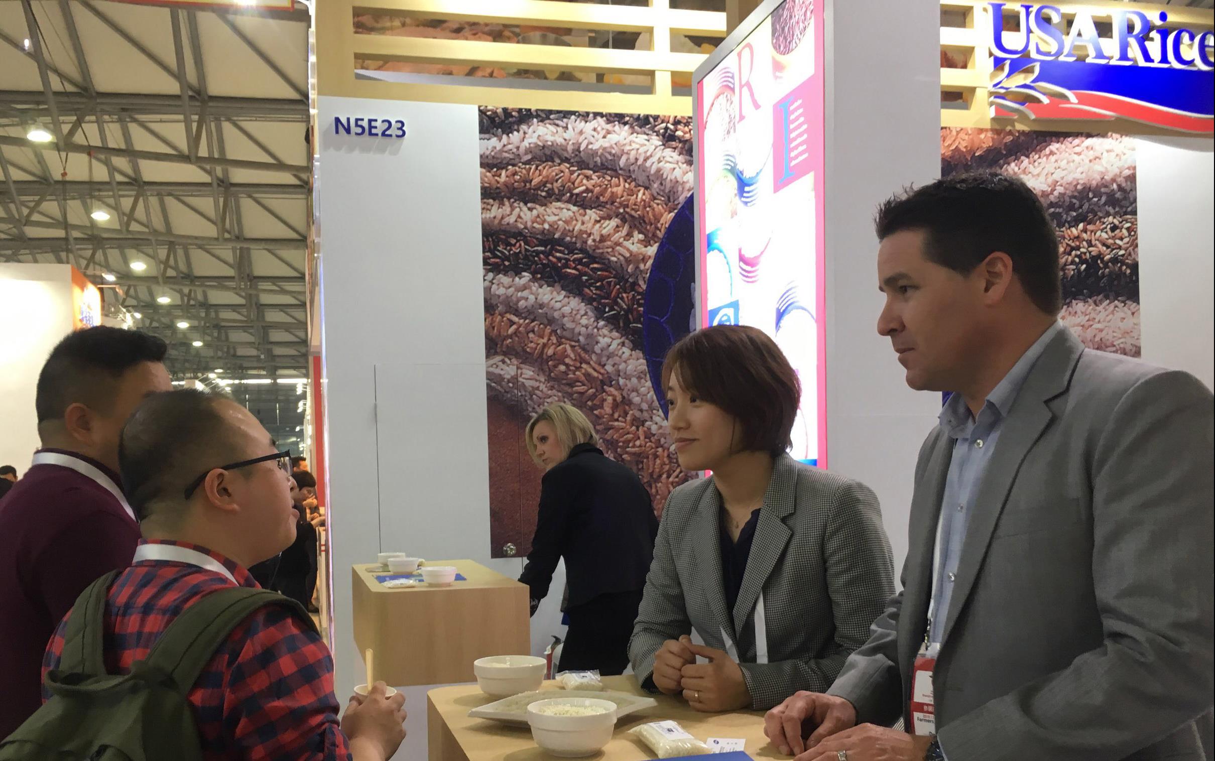 People gather at a trade show booth, standing around a table filled with rice bowls and pamphlets