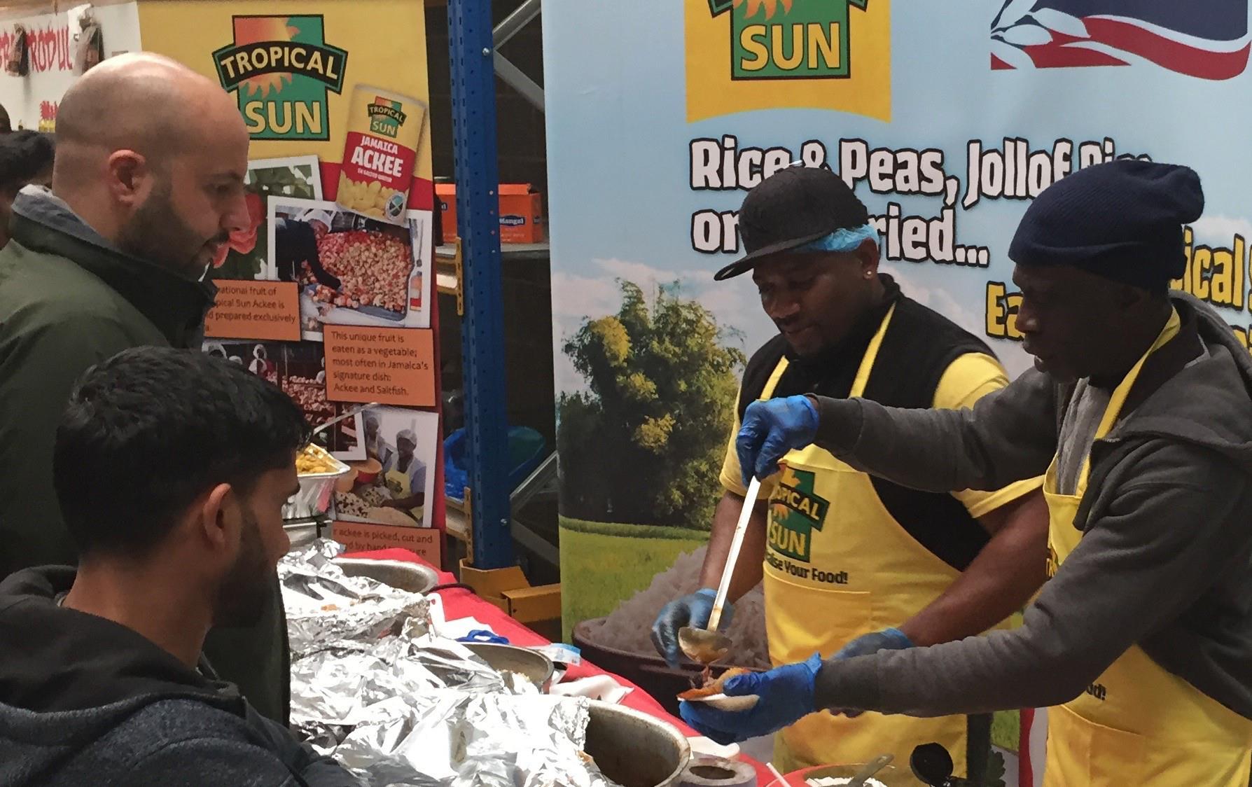 People gather at a buffet table filled with colorful dishes being served by two black men wearing yellow aprons standing in front of a USA Rice & Tropical Sun banner