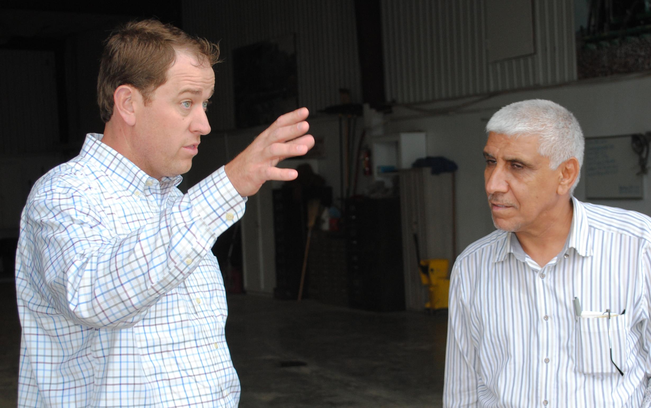 Two men wearing long-sleeved button-down shirts meet inside an equipment shed, one on the left gesturing with one hand, one on the right listens intently