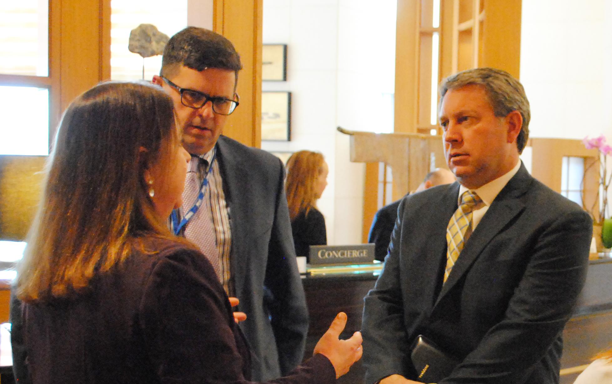 A woman and two men wearing business suits stand talking in front of hotel desk