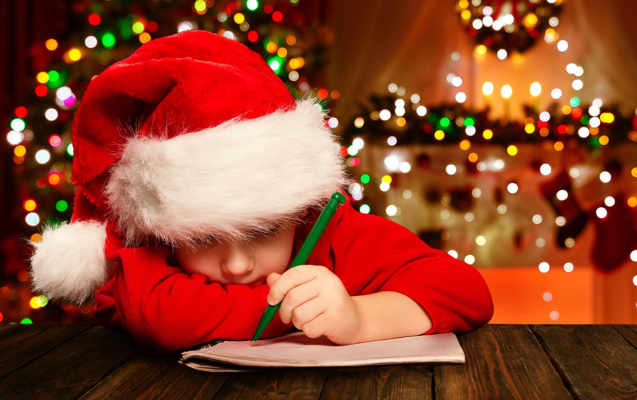 Child wearing Santa hat sits at a table writing on a tablet, Christmas lights in the background