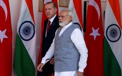 Two older men walk in front of flags of Turkey and India, one wears a business suit, the other wears a white shirt and light blue vest