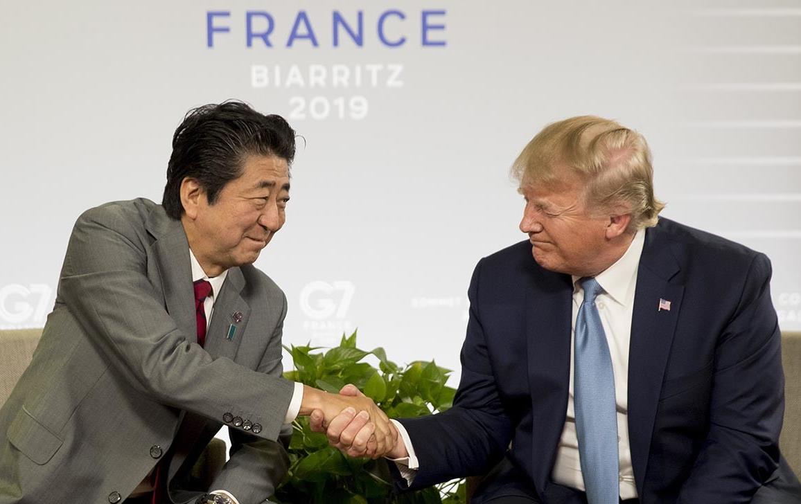 Japanese Prime Minister Shinzo Abe shakes hands with US President Donald Trump, both men wearing business suits, seated in front of backdrop reading Biarritz 2019