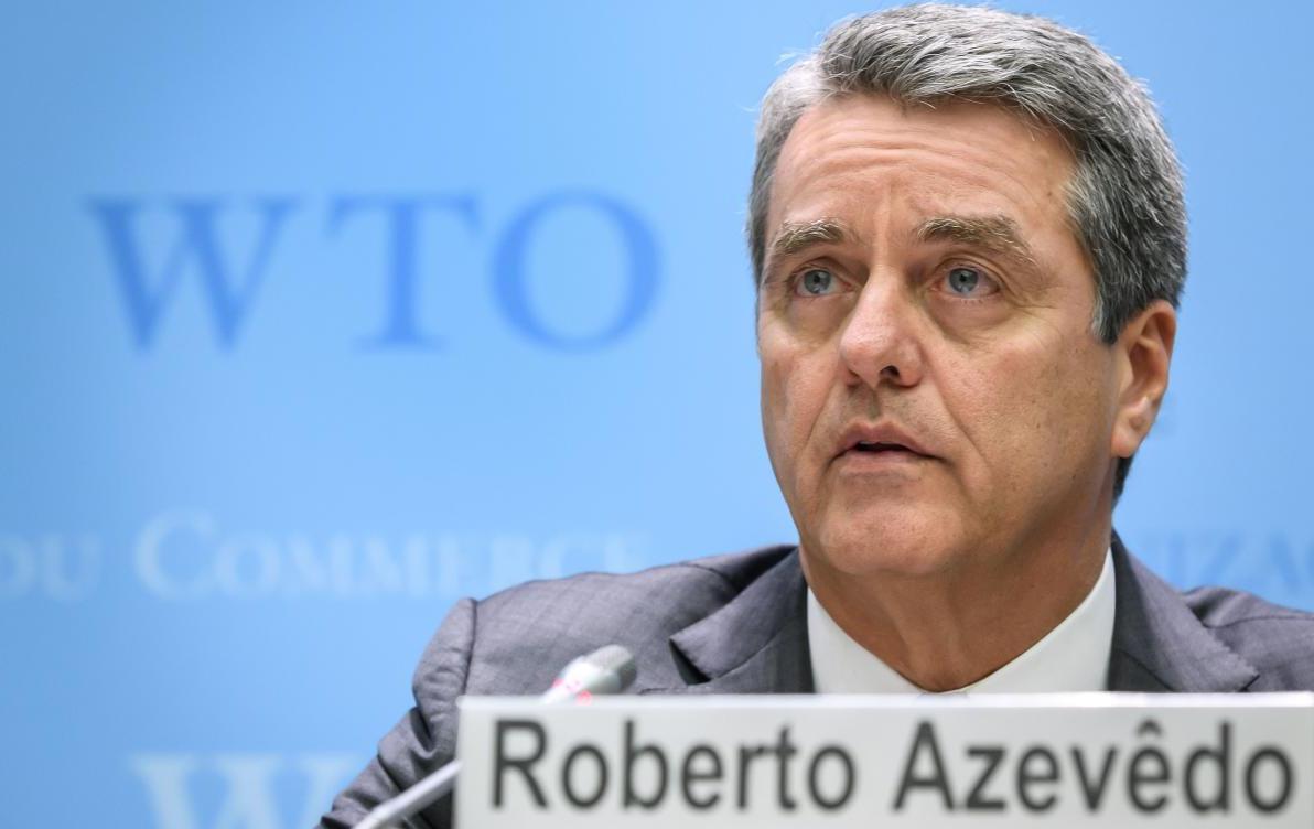 WTO Director General Roberto Azevedo sits at table with name plate and "WTO" on wall in background