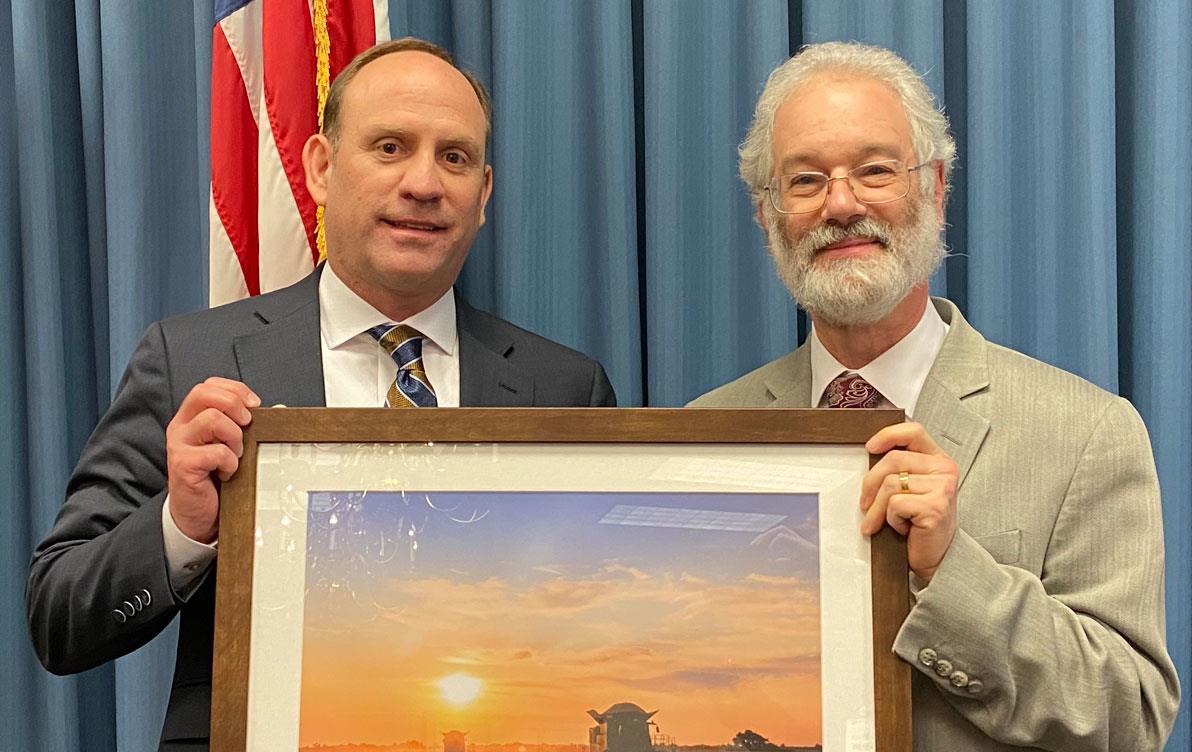 Two men wearing business suits hold framed photo of combines in a rice field; blue curtain background with U.S. flag