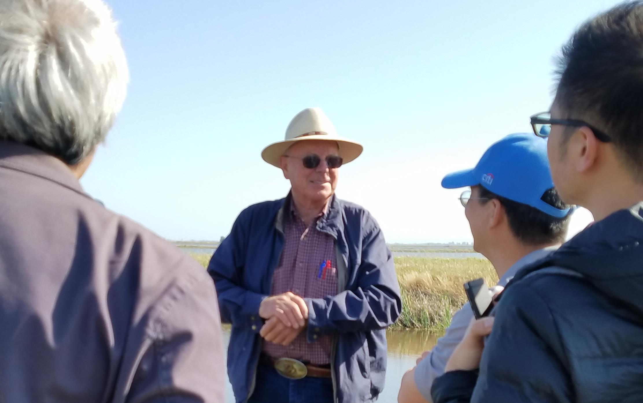 Farmer wearing straw hat and sunglasses stands in middle of group of people listening to his lecture, rice field and drainage ditch in background