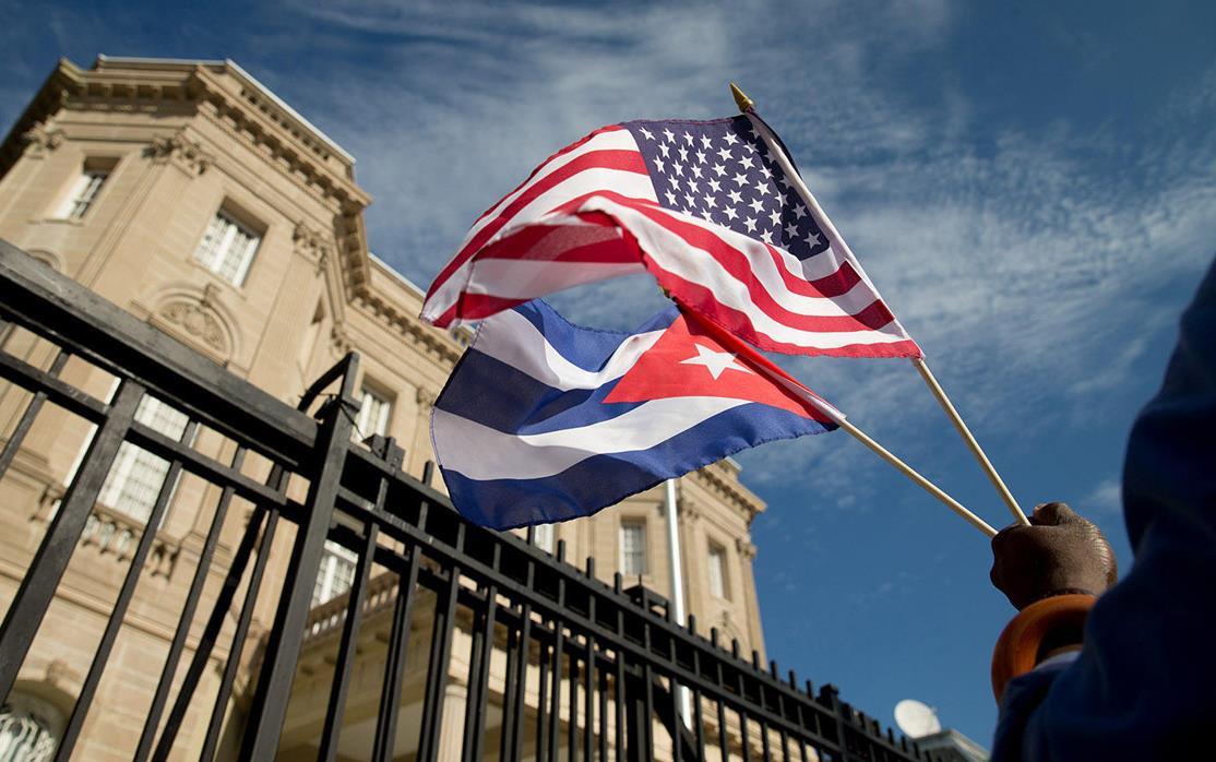 Hand holds US and Cuban flags in front of embassy building and fence