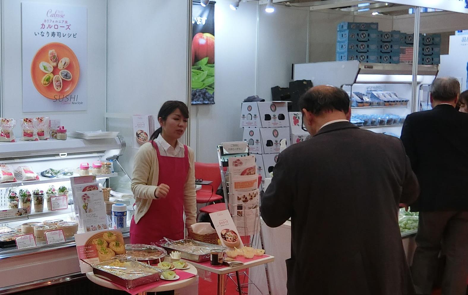 Trade show booth with people standing around talking, a woman wearing a red apron offers food samples to man wearing black suit