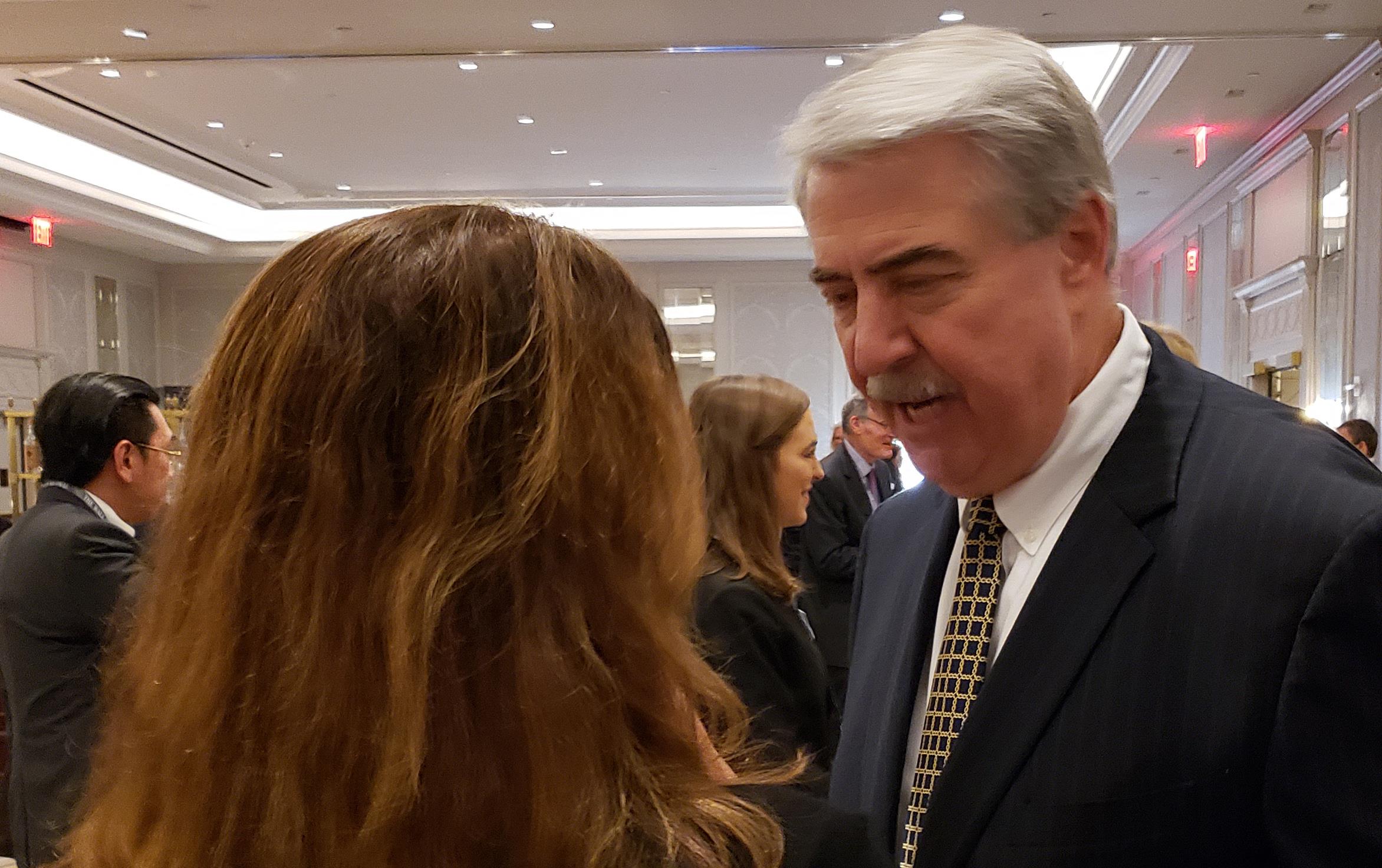 Woman with dark hair and back to camera talks with older white man with mustache wearing business suit