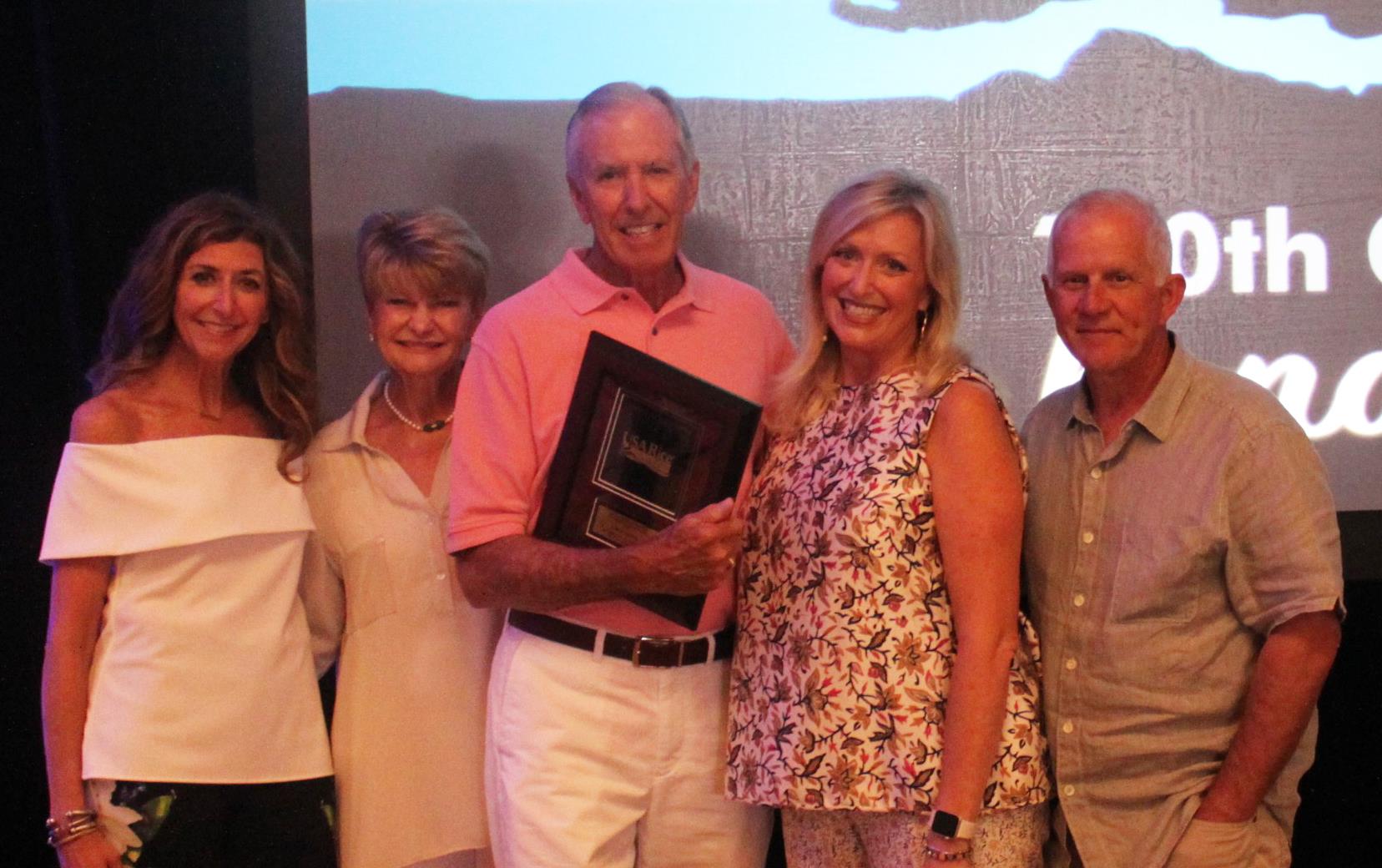 Family group dressed in casual clothes stands around central older white male who holds commemorative plaque