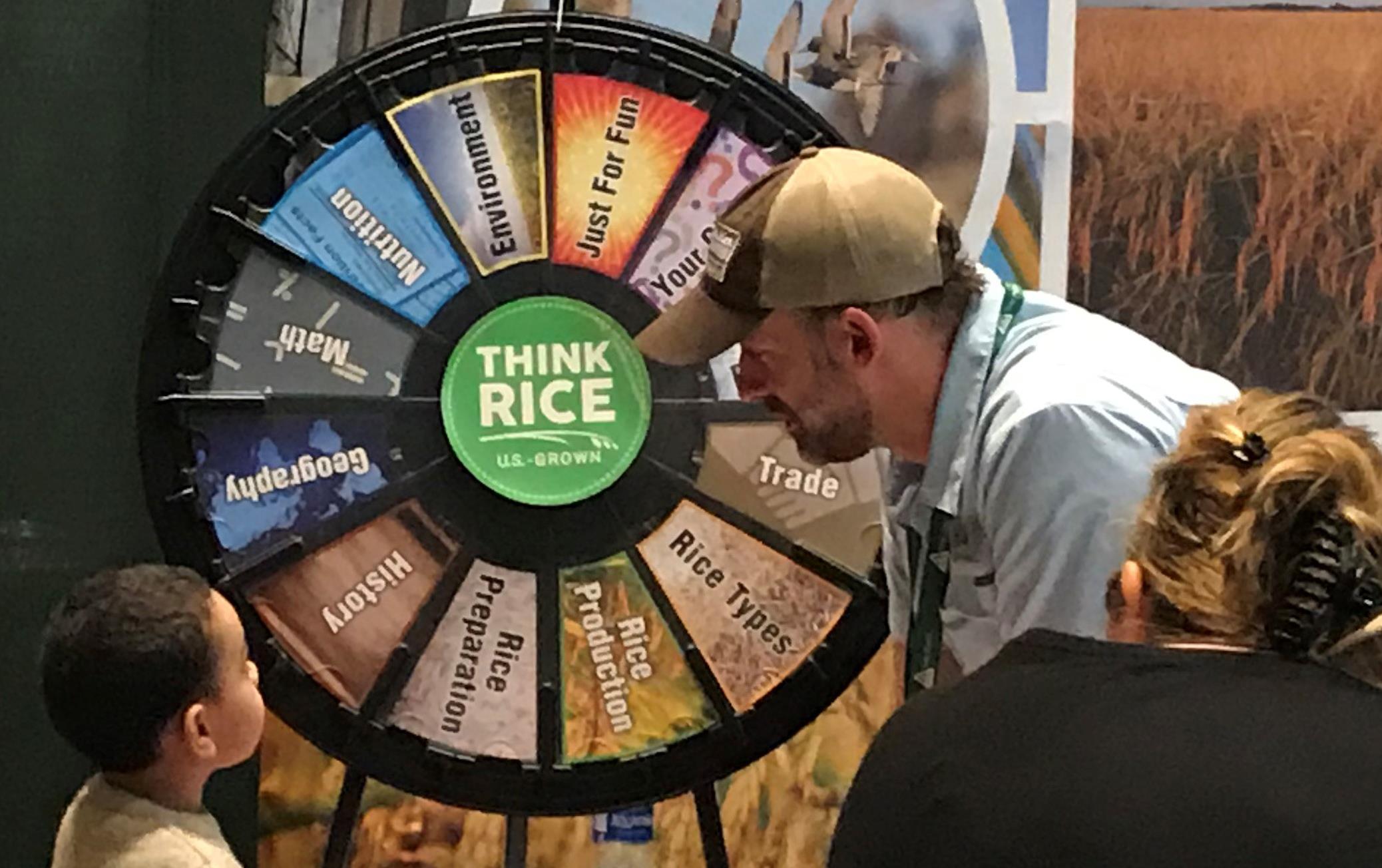 Children line up for their chance at the Think Rice trivia wheel sitting on a table draped in an American flag tablecloth, man wearing ballcap leans over to talk to small boy getting ready to spin the wheel