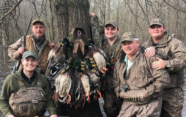 Group of men dressed in camoflage standing in knee-deep muddy water with a brace of dead ducks hanging from a tree