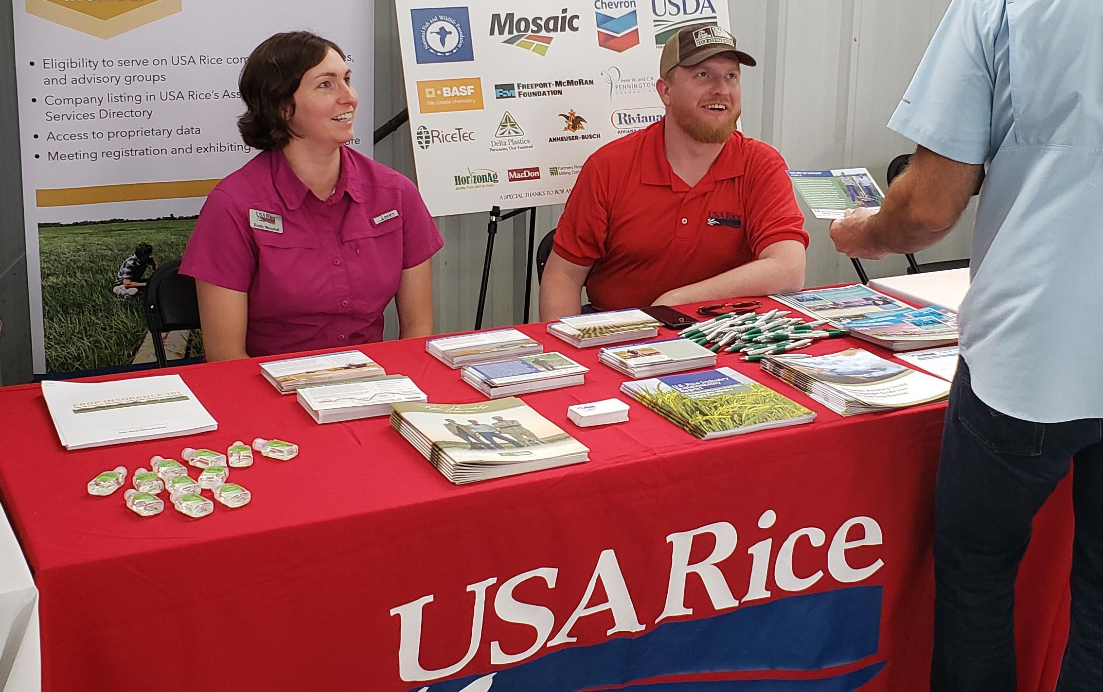 A woman and a man sit behind a table draped with USA Rice tablecloth and covered with literature, man stands in front holding postcard, posters in background