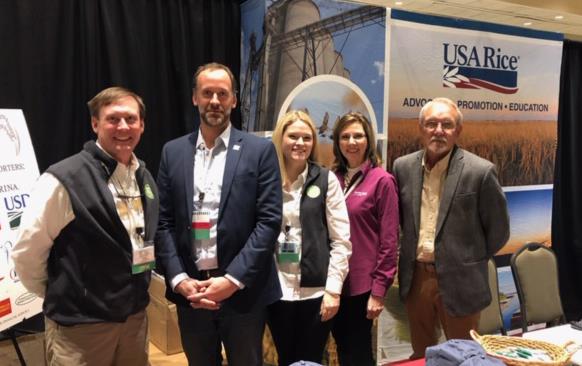 Group shot, men and women standing in USA Rice booth behind table filled with ballcaps, pens, and brochures