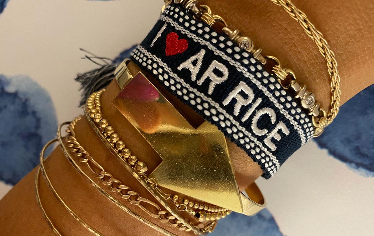 Close-up of I Heart AR Rice bracelet worn w/other gold bracelets, arrow pointing down