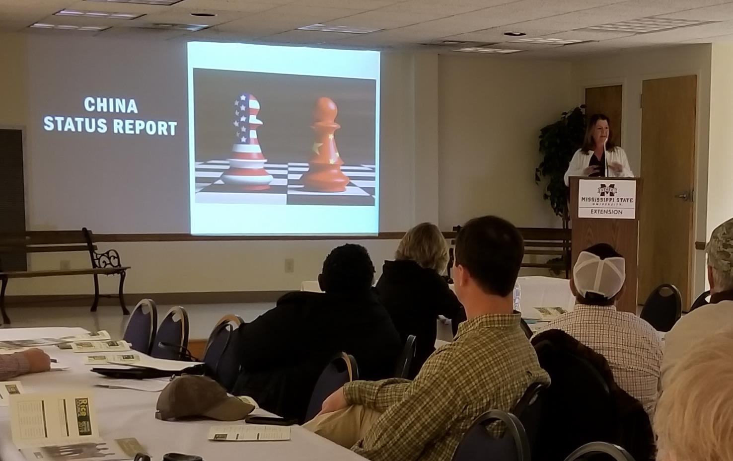 Woman talks from podium, standing beside projection screen, audience sits in folding chairs at long tables