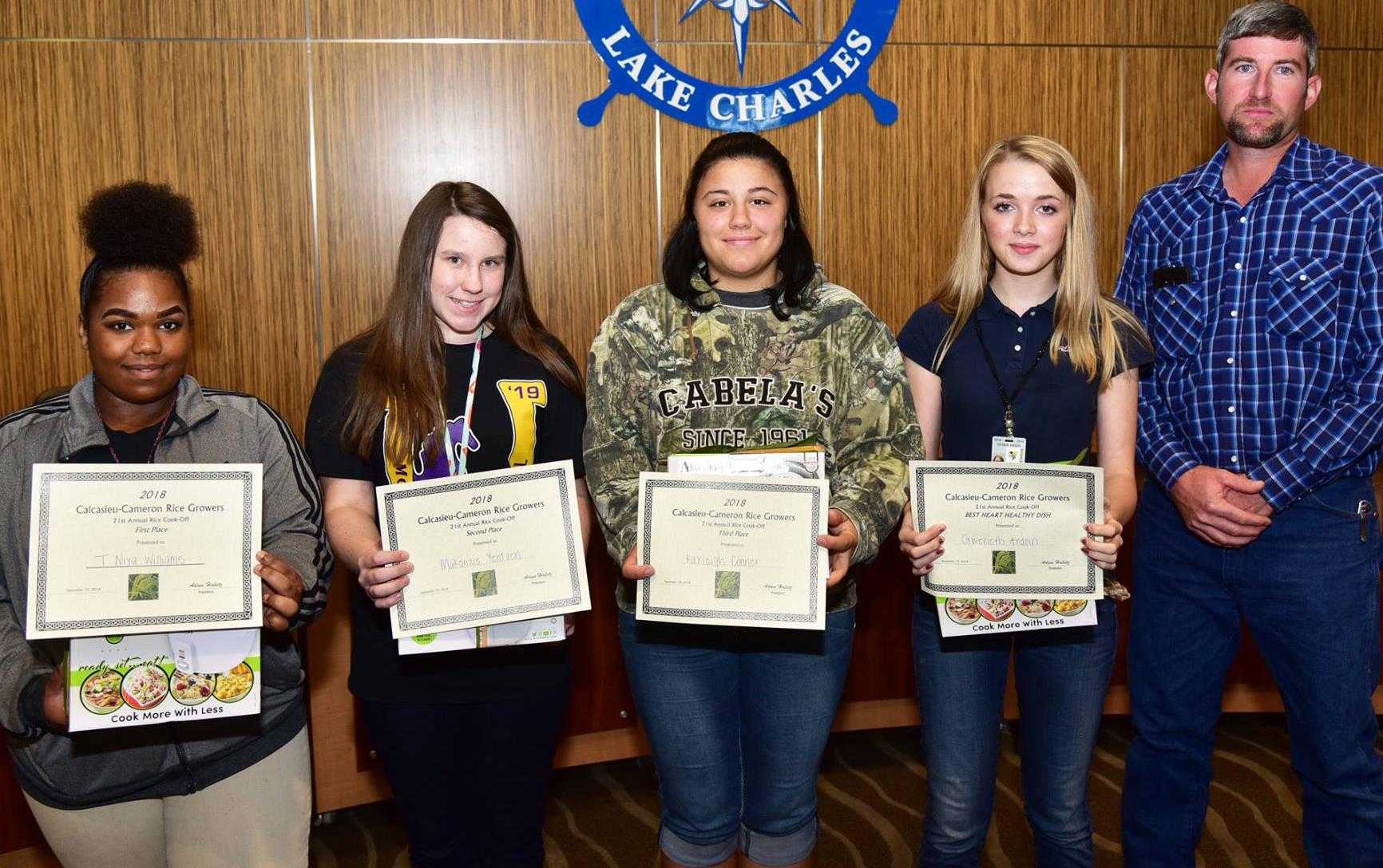 Four female students holding certificates and rice cookers stand in front of Port of Lake Charles logo, male teacher wearing blue plaid shirt stands on the far right