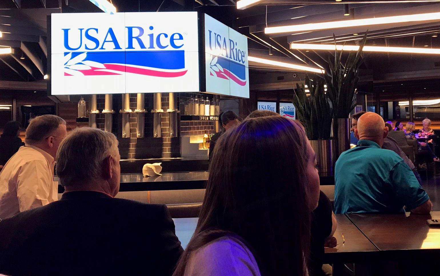 Group of people seated with backs to the camera in a large meeting room, USA Rice text and logo illuminated on big screens