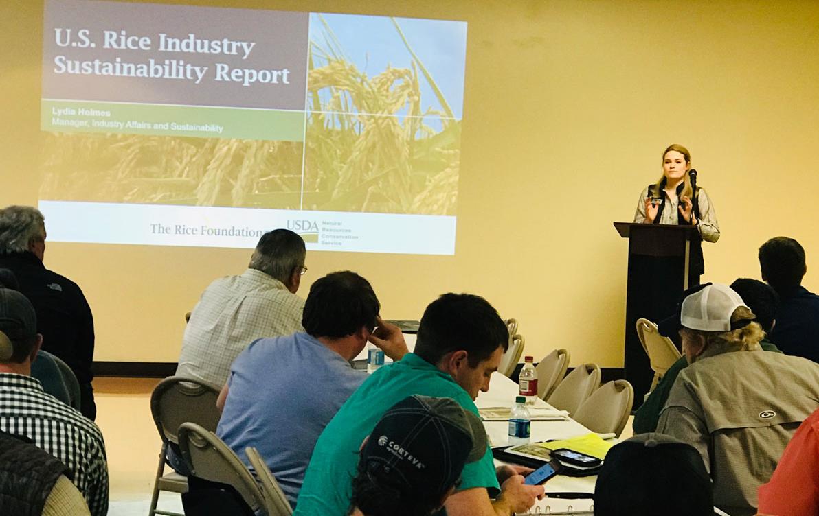 Young woman stands at podium delivering slide presentation to crowded room of people seated at tables