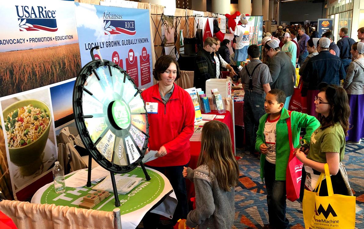 Woman wearing red shirt stands in exhibit booth in front of children gathered around spinning Rice Wheel