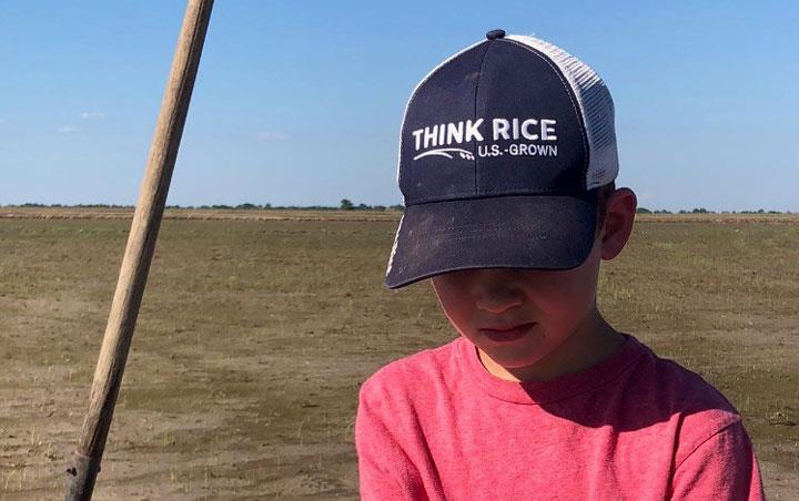 Young boy wearing jeans, red shirt, and ThinkRice ballcap kneeling in planted field, holding new rice plant