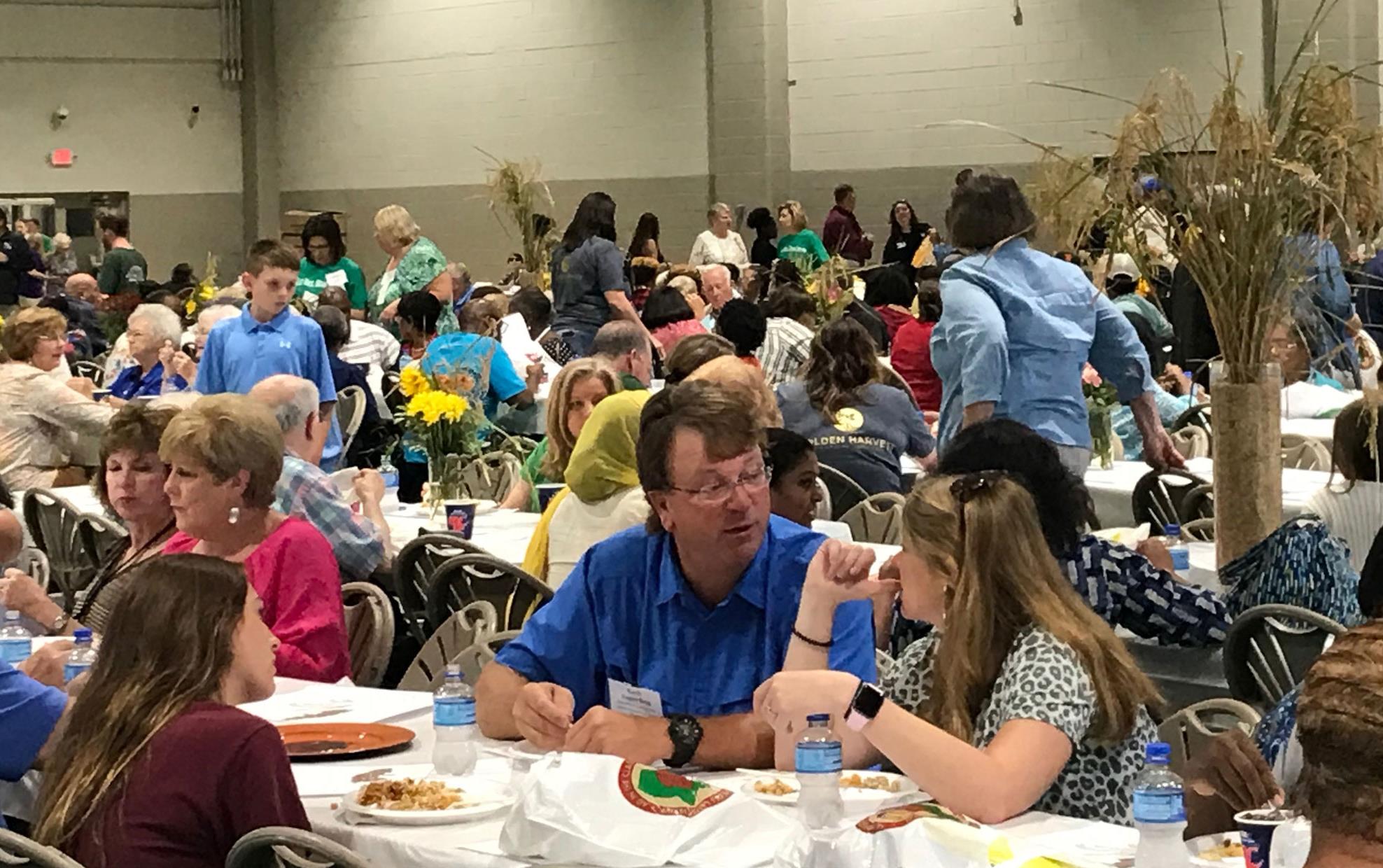 Crowds of people seated at tables enjoying plates of food, rice stalks as centerpieces