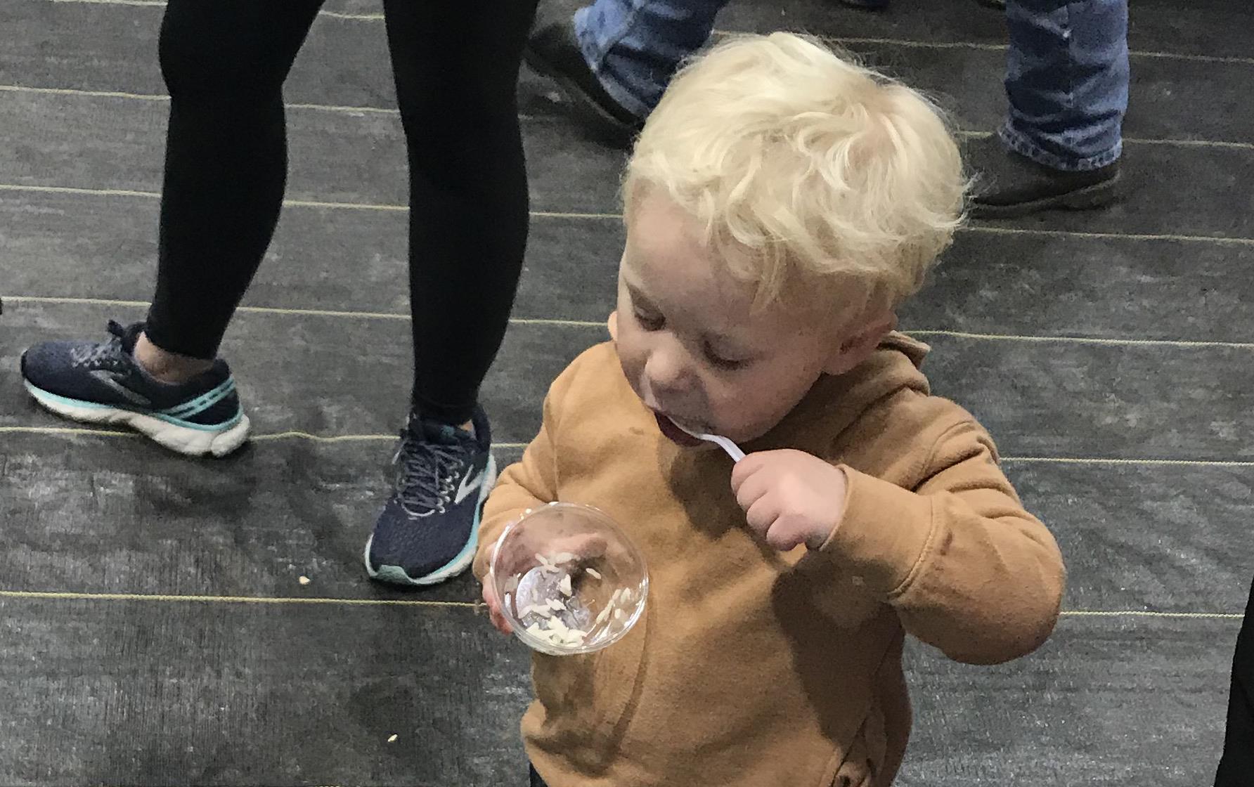 Young, blond boy eats rice out of plastic cup in front of table filled with bags of jasmine rice